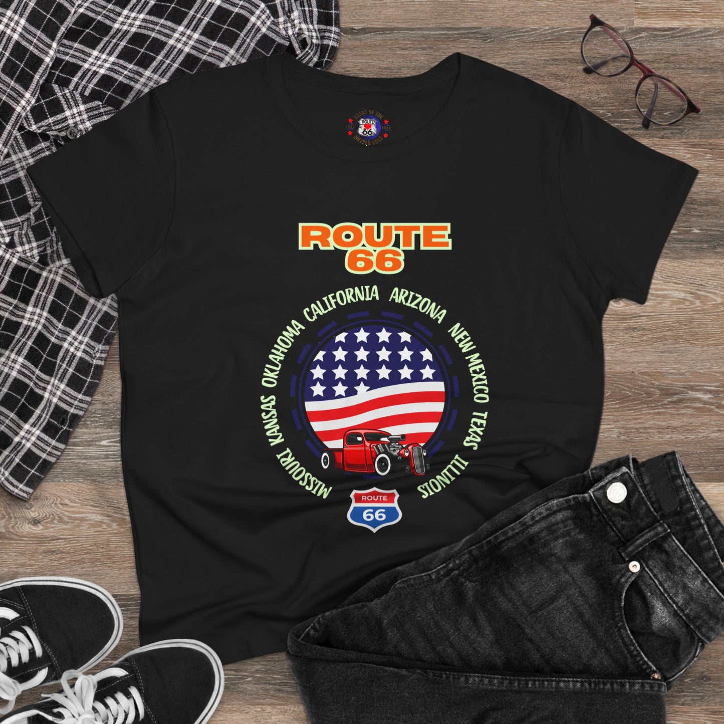 A black Route 66 t-shirt is laid flat on a wooden surface, surrounded by black jeans, plaid fabric, sneakers and glasses. The shirt features a graphic with "ROUTE 66" text and a large circular American flag design. Inside the circle is a red classic hot rod car. Around the flag are the states written: Missouri, Kansas, Oklahoma, Texas, New Mexico, Arizona, California, and Illinois.