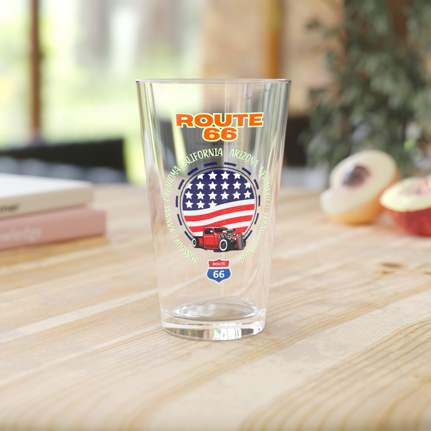 Front view of Route 66 16oz pint glass on top of a wooden kitchen counter. The design features a patriotic American flag circle, a vintage red truck graphic, and all eight states of the Mother Road circling the design