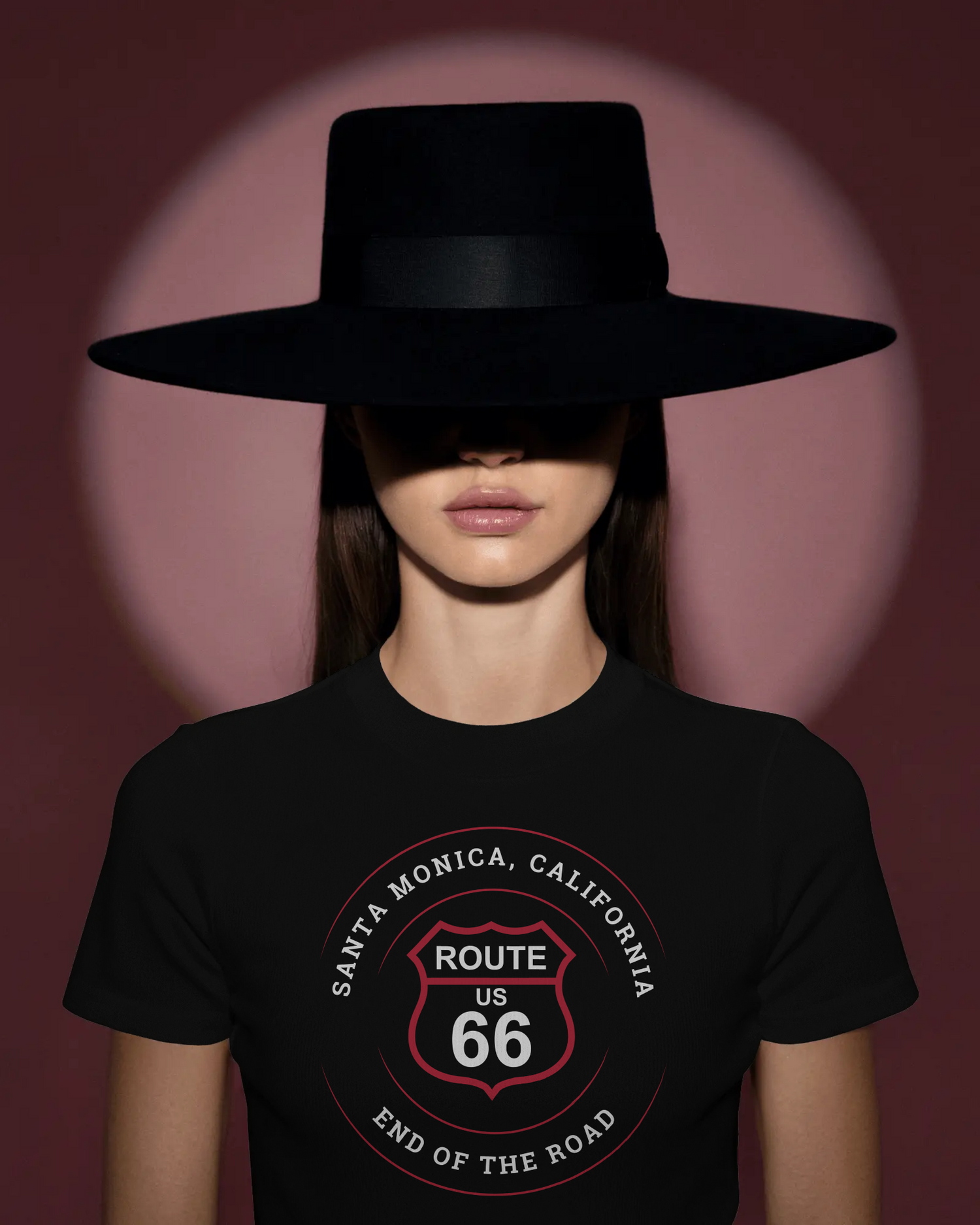 Woman wearing a wide-brimmed black hat and a Route 66 Santa Monica "End of the Road" t-shirt, standing in dramatic lighting.