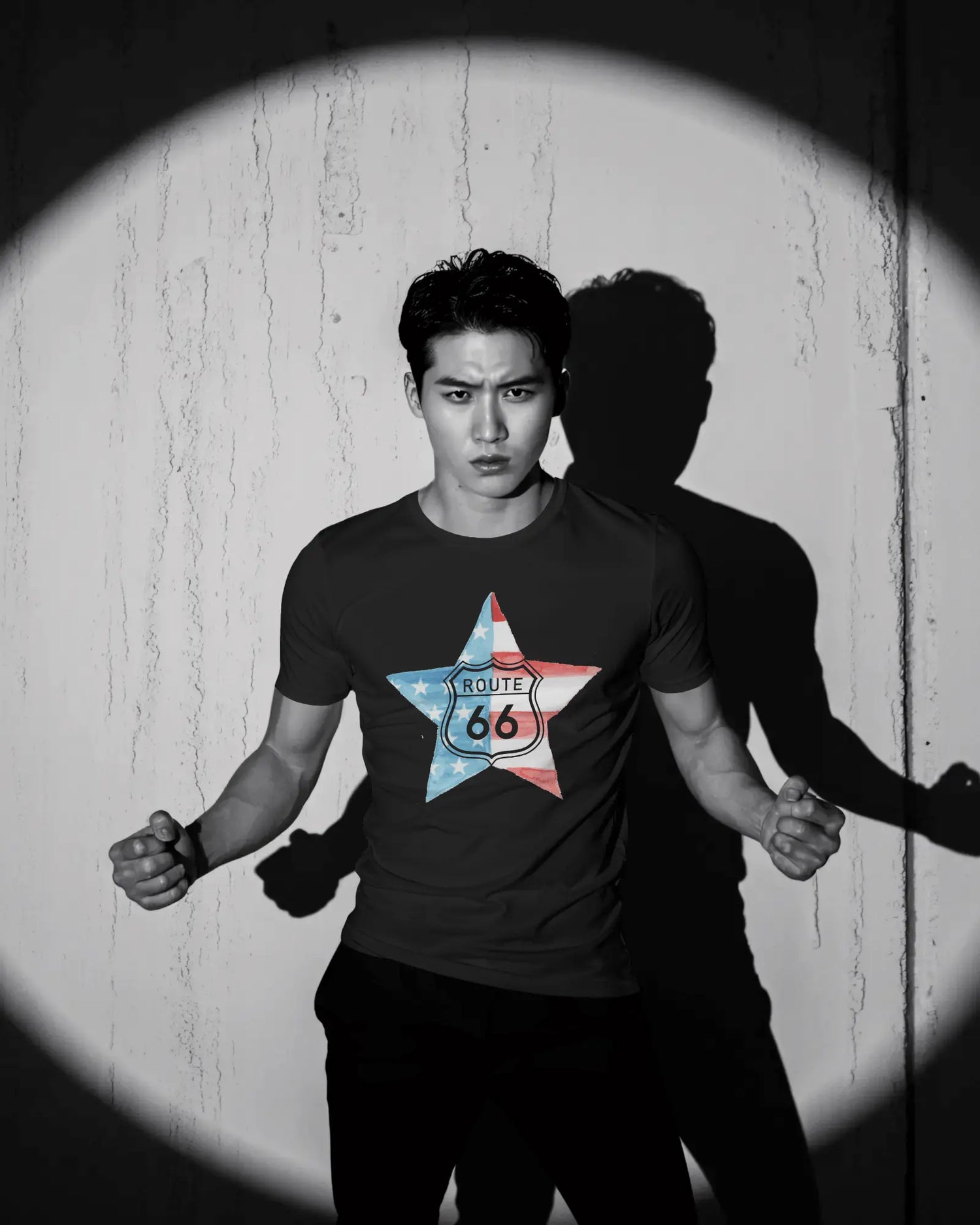 A black and white photo shows a man in a t-shirt with a Route 66 star logo, standing under a spotlight with his shadow cast behind him. He has a serious expression and his hands are clenched.