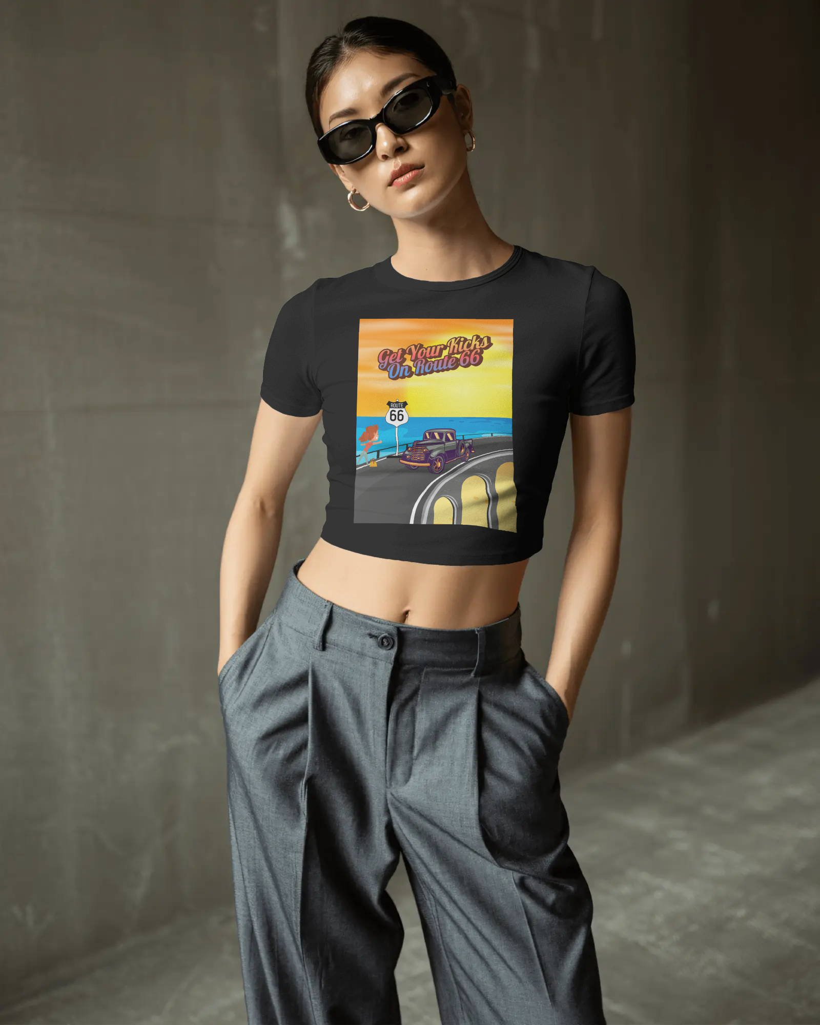 Woman wearing a back retro Route 66 women's baby tee with colorful "Get Your Kicks on Route 66" graphic, styled with sunglasses and high-waisted pants.