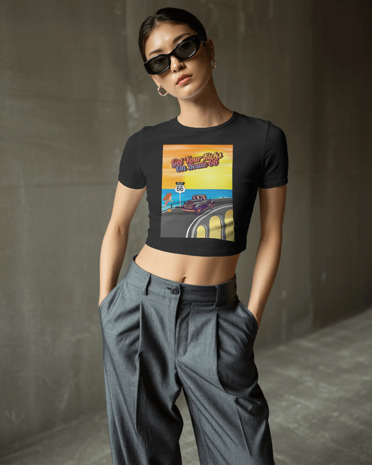 Woman wearing a back retro Route 66 women's baby tee with colorful "Get Your Kicks on Route 66" graphic, styled with sunglasses and high-waisted pants.