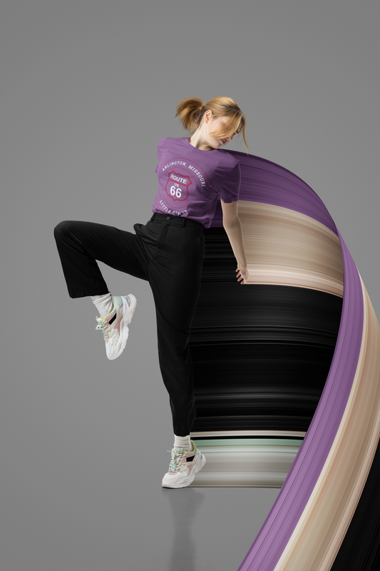 A dynamic full-body studio shot of a person from the back, wearing a heather team purple retro Route 66 unsiex jersey tee with an Arlington, MO: "Little Piney" design. She also has black pants and white sneakers with colorful accents. The woman is mid-action, with one leg bent, and a purple, black, and beige ribbon-like fabric sweeps out to their right. 