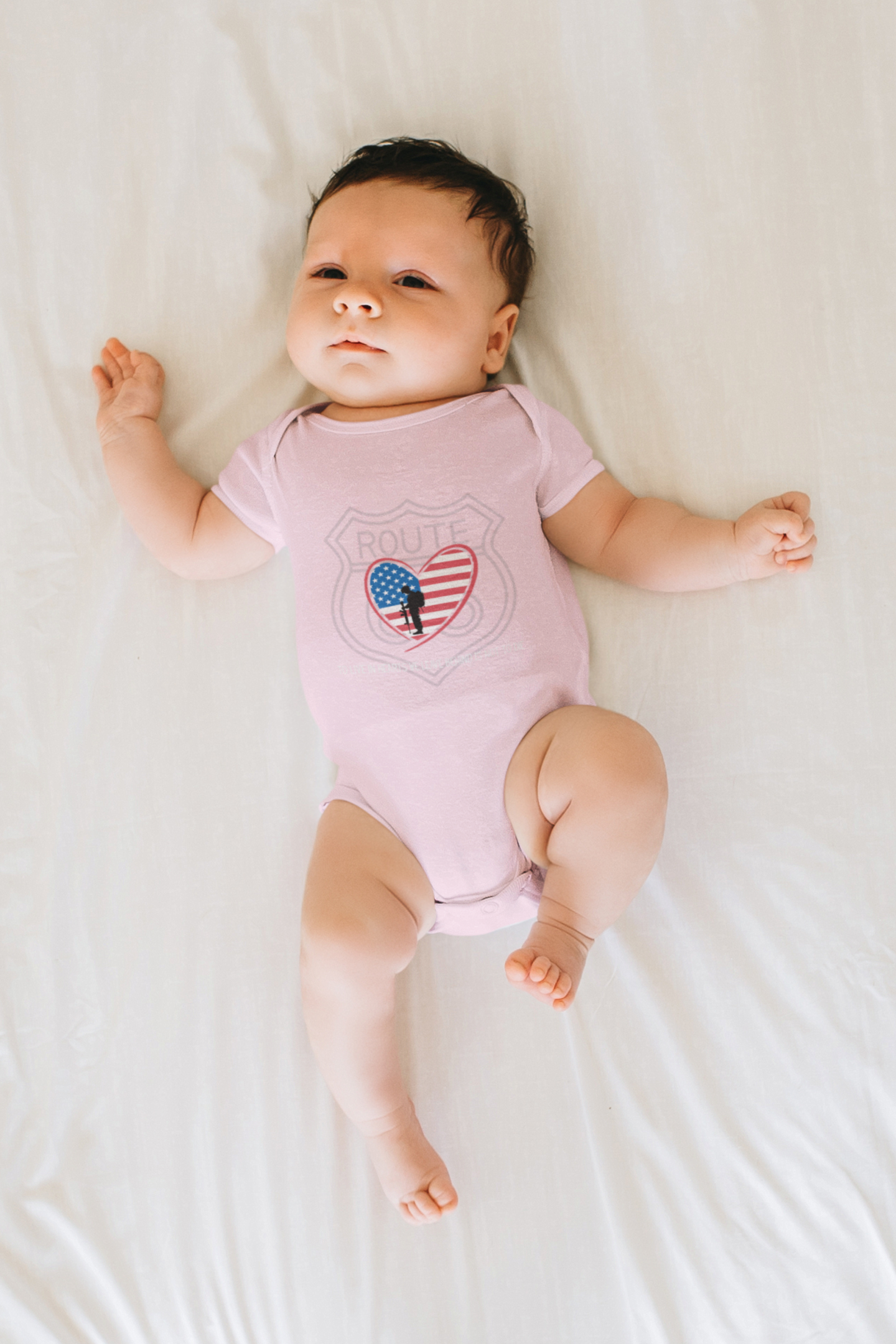 Baby lying on a white sheet and wearing a pink Route 66 patriotic heart onesie featuring an American flag heart and fallen soldier silhouette.