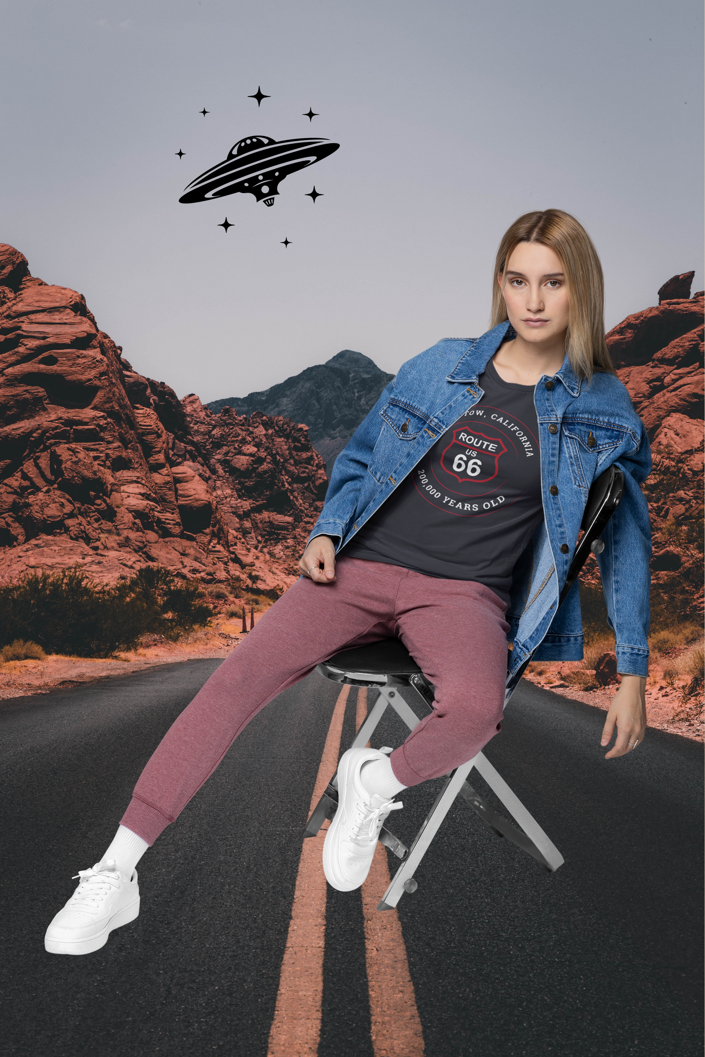 Female model sitting on a chair in the middle of Route 66 with a UFO flying above and wearing a vintage black retro Route 66 unisex jersey tee with Barstow, CA: "200,000 Years Old" design