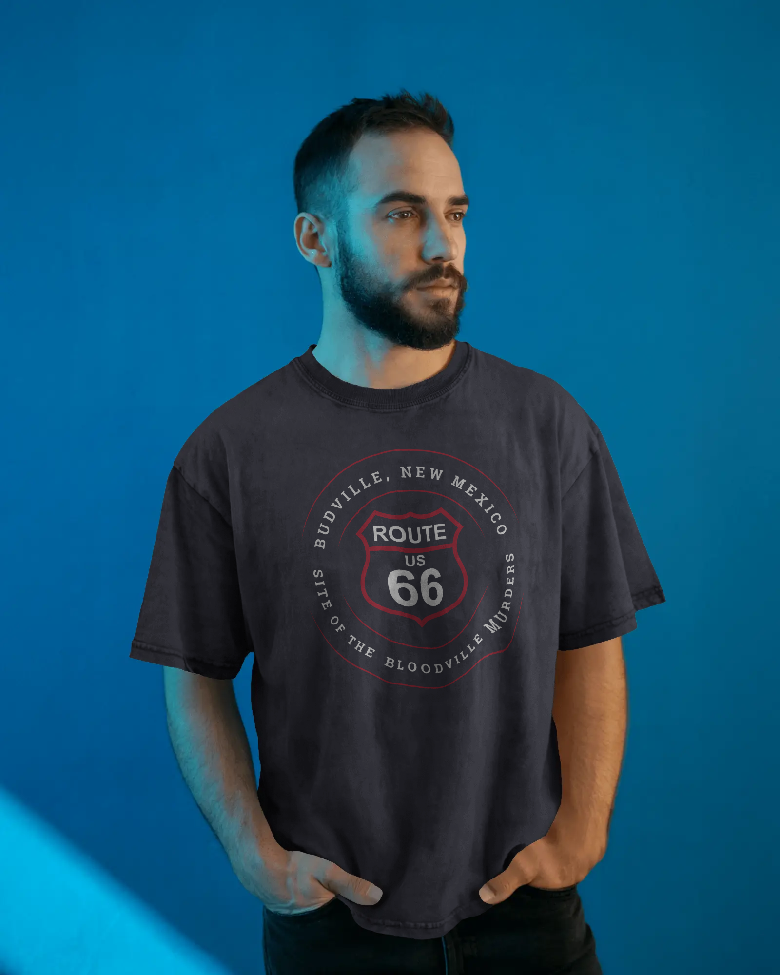 Male model wearing dark grey heather retro Route 66 unisex jersey tee with Budville, NM: Site of the Bloodville Murders graphic
