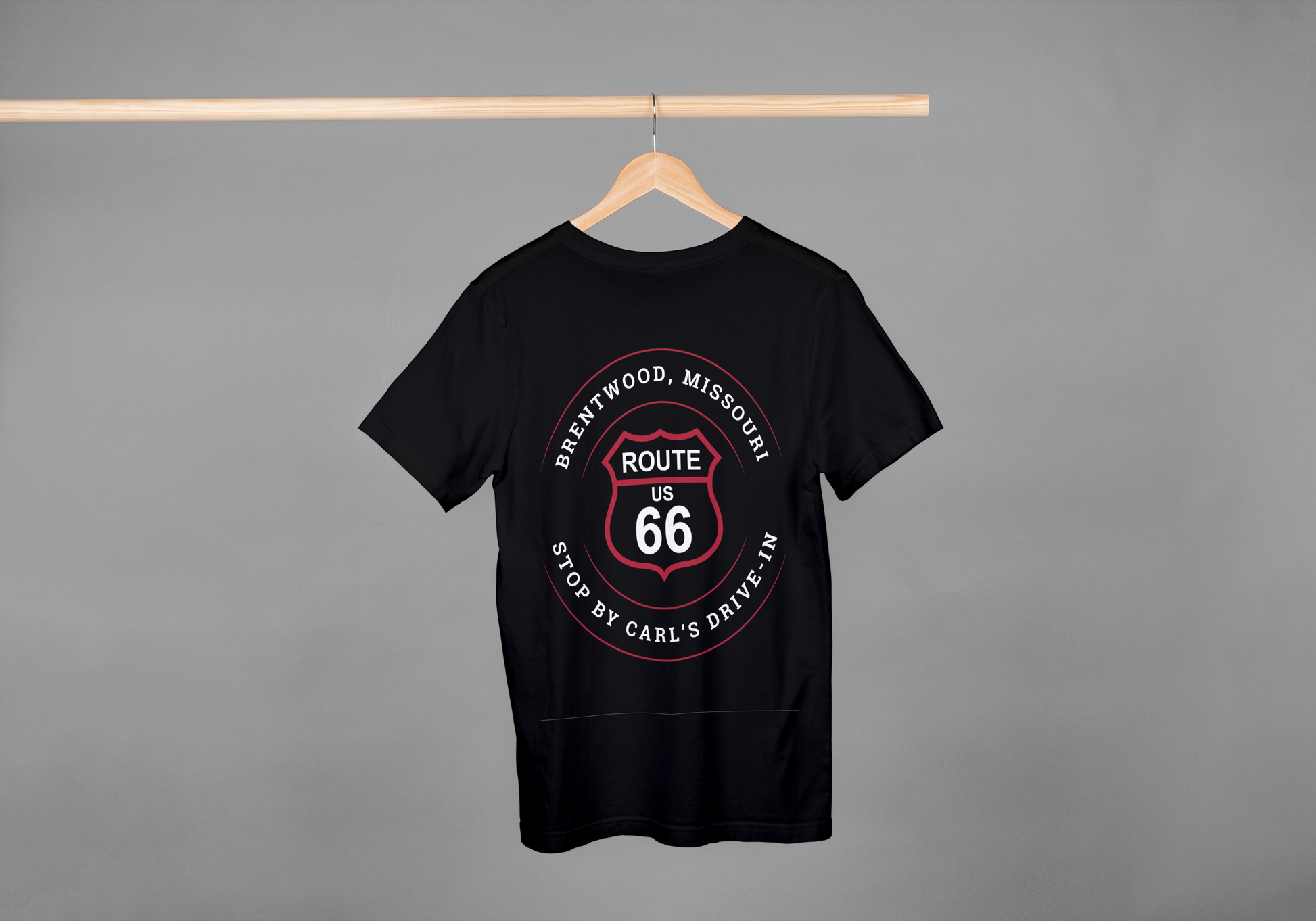 A black retro Route 66 unisex jersey tee with a Brentwood, MO: "Stop By Carl's Drive-In" design hanging off a wooden hanger on a pole