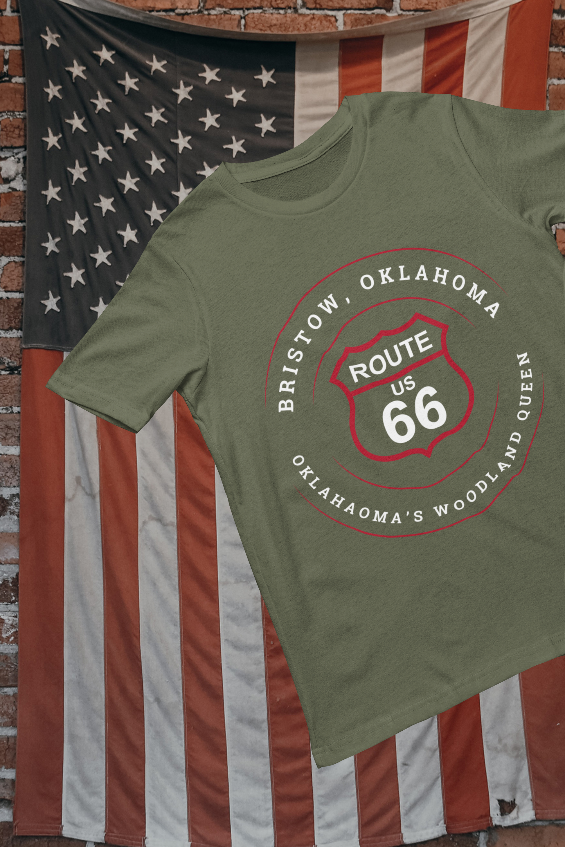 Heather military green retro Route 66 unisex tee with Bristow, OK 'Oklahoma's Woodland Queen' graphic displayed against a weathered American flag backdrop on a brick wall.