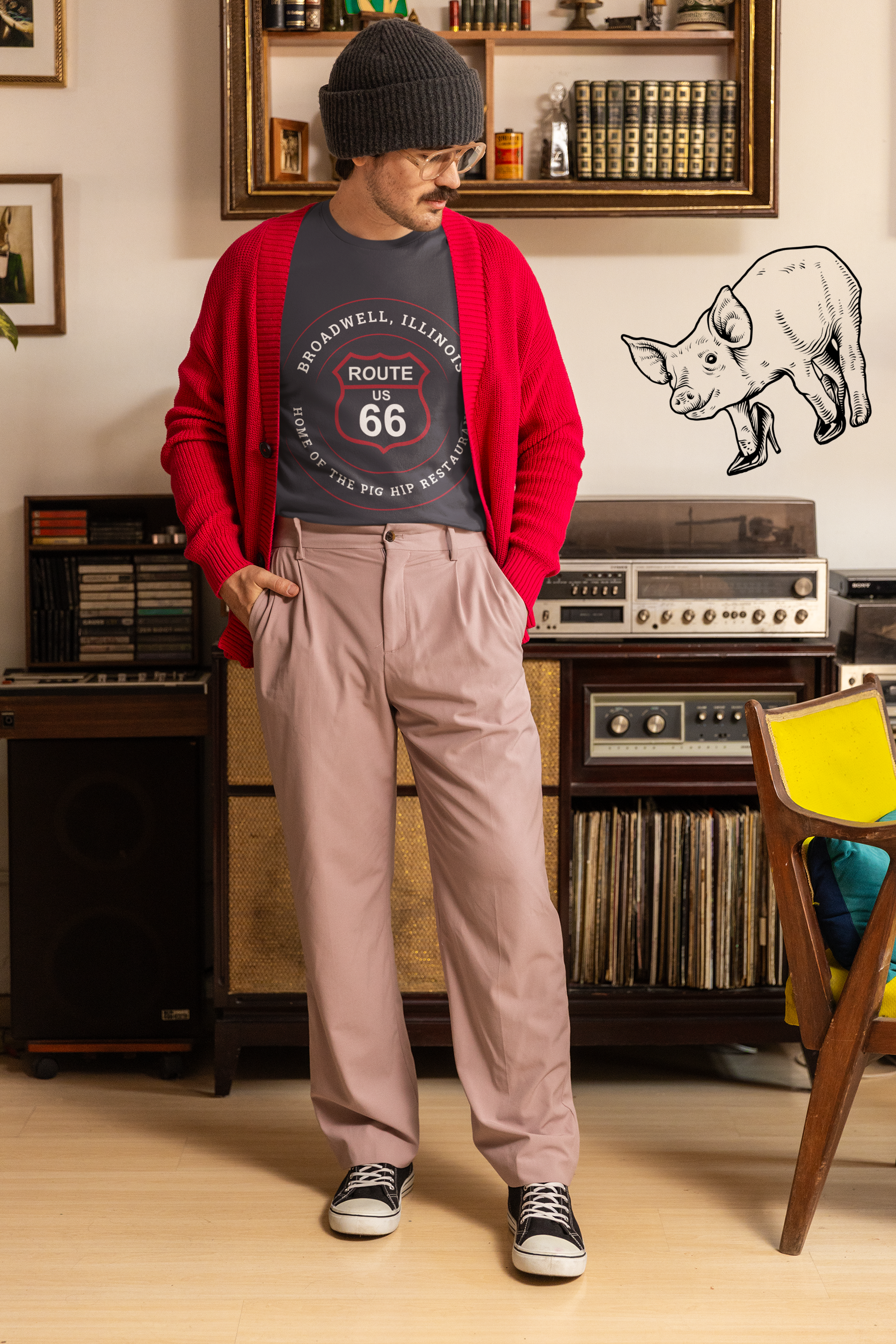 Female model wearing a dark grey heather retro Route 66 unisex jersey tee with a Broadwell, IL: "Home of the Pig Hip Restaurant" design