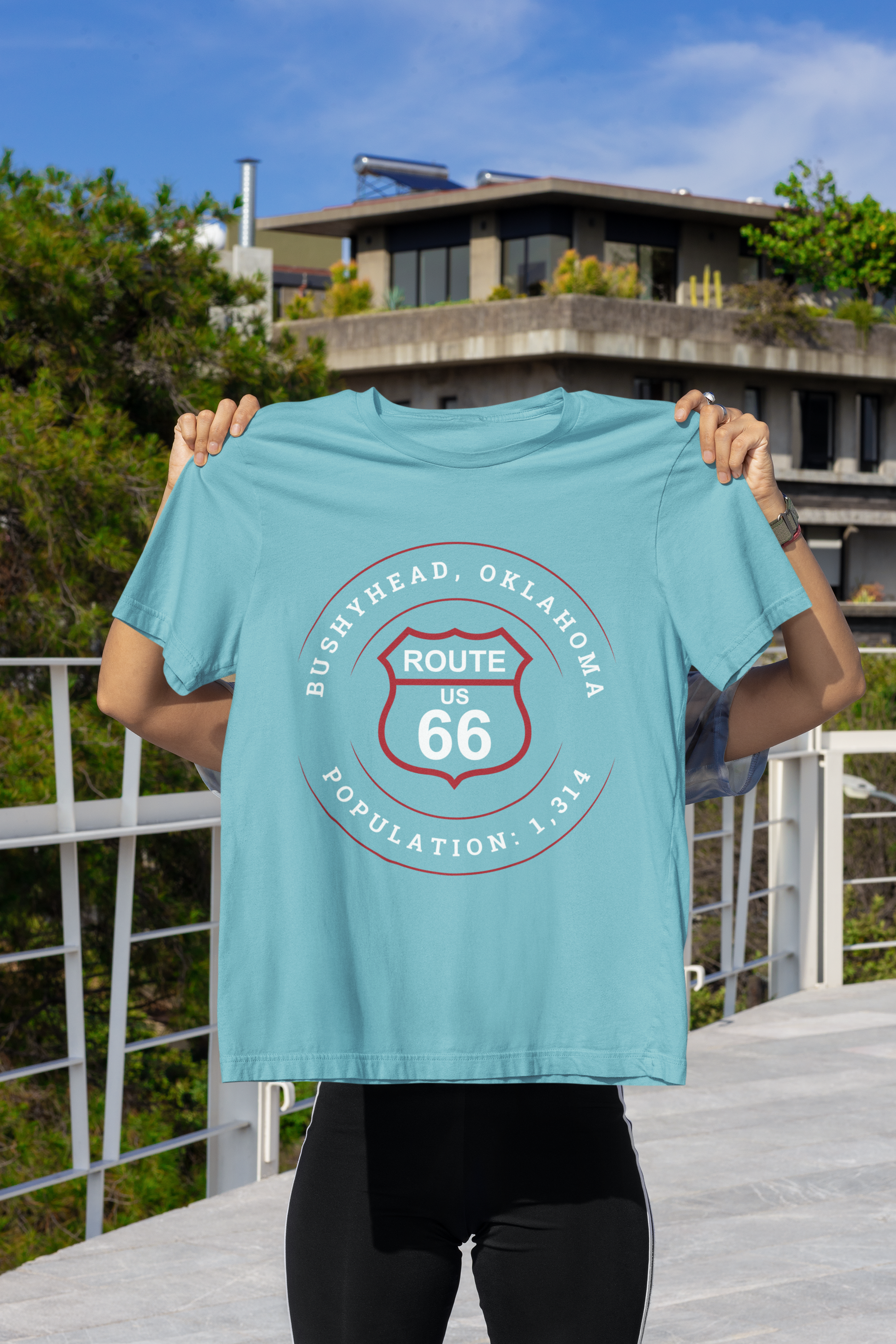 Turquoise retro Route 66 unisex jersey tee with Bushyhead, OK "Population 1,314"  design being held up by a female at an apartment complex