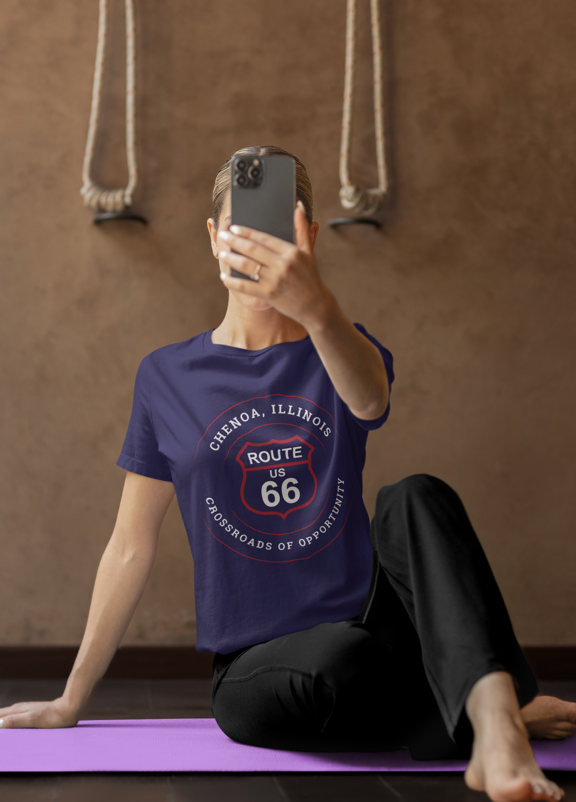 Female model taking a selfie with her mobile phone, and she is wearing a team navy retro Route 66 unisex jersey tee with a Chenoa, IL: "Crossroads of Opportunity" design