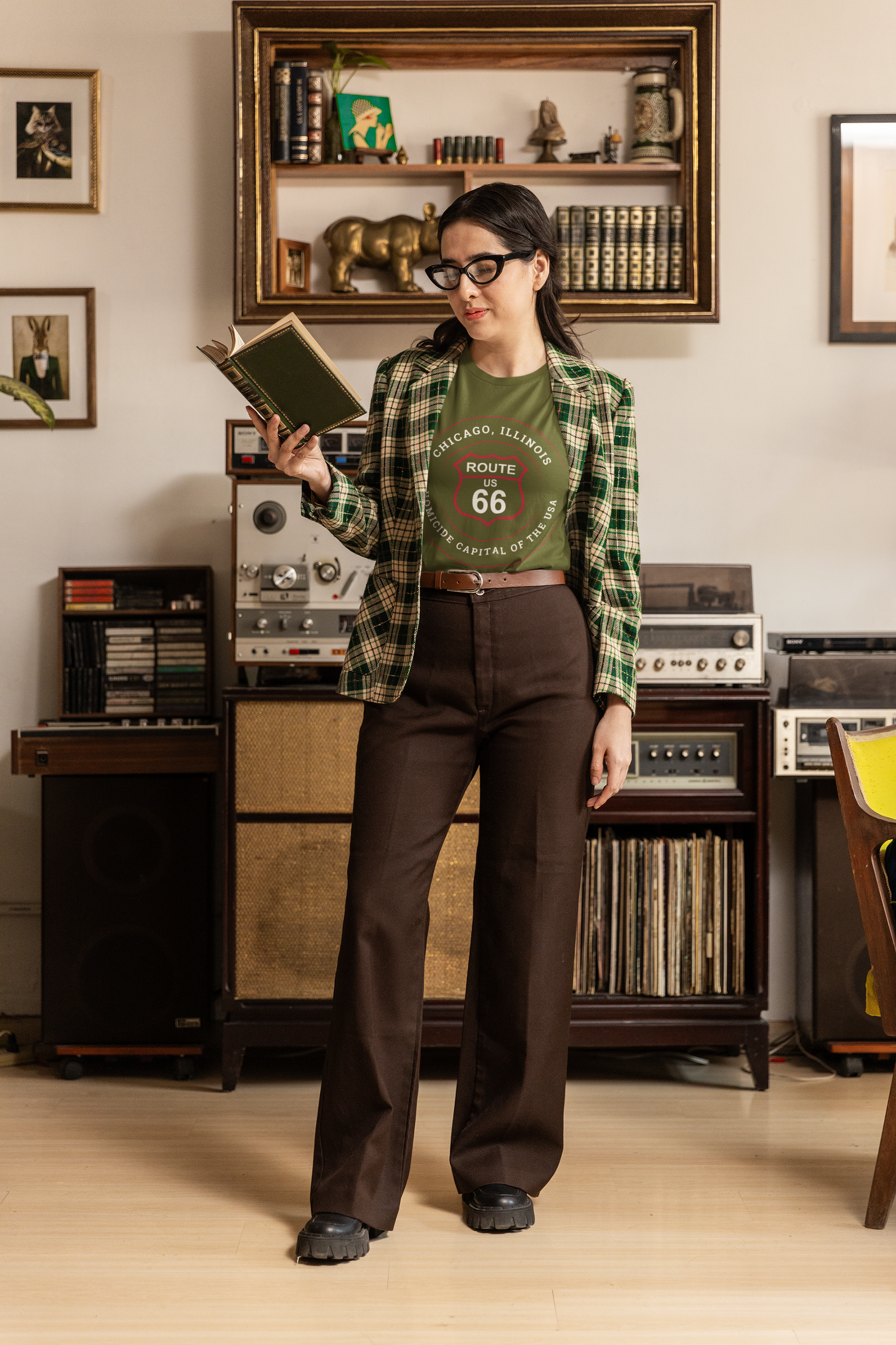 Female model is reading a book and wearing a heather military green retro Route 66 unisex jersey tee with a Chicago, IL:: "Homicide Capital of the USA" design, along with a green and white jacket