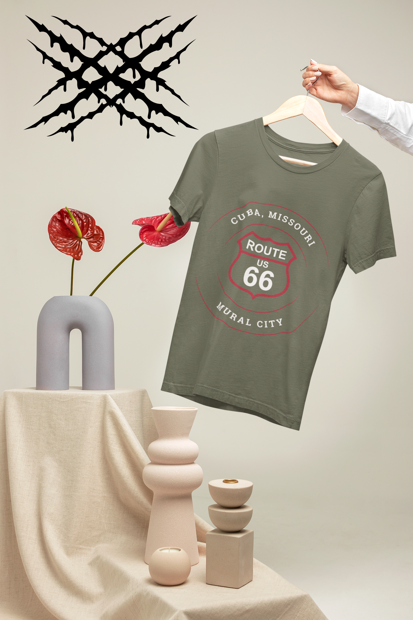 Person's arm is in the photo and is holding a heather military green retro Route 66 unisex jersey tee with a Cuba, MO: "Mural City" design on a hanger