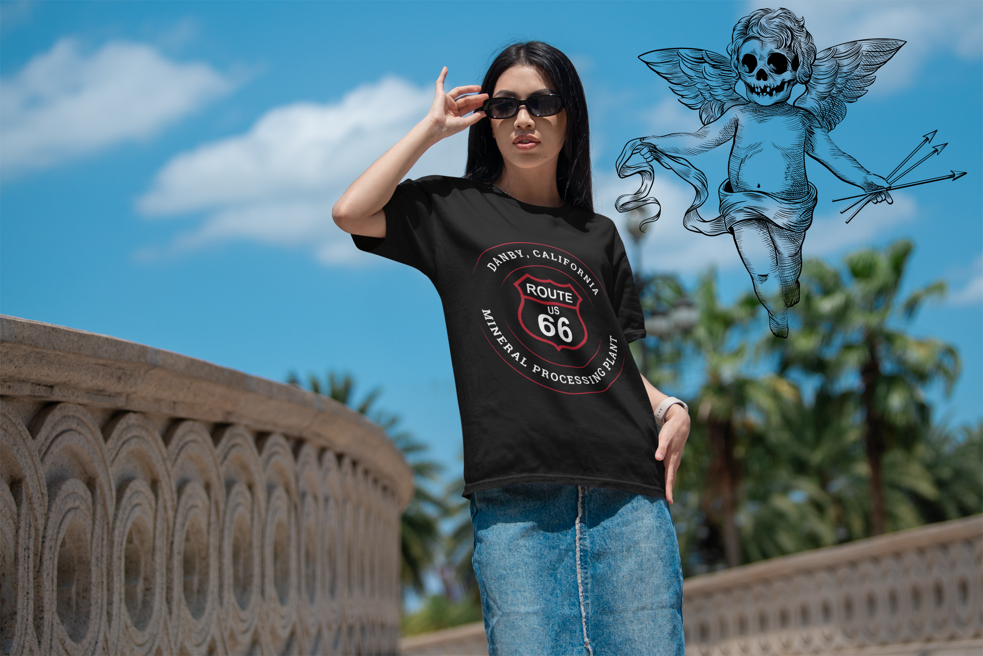 Female model wearing black retro Route 66 unisex jersey tee with Danby, CA: "Mineral Processing Plant" design with a blue sky background and an angel with a skullface holding three arrows