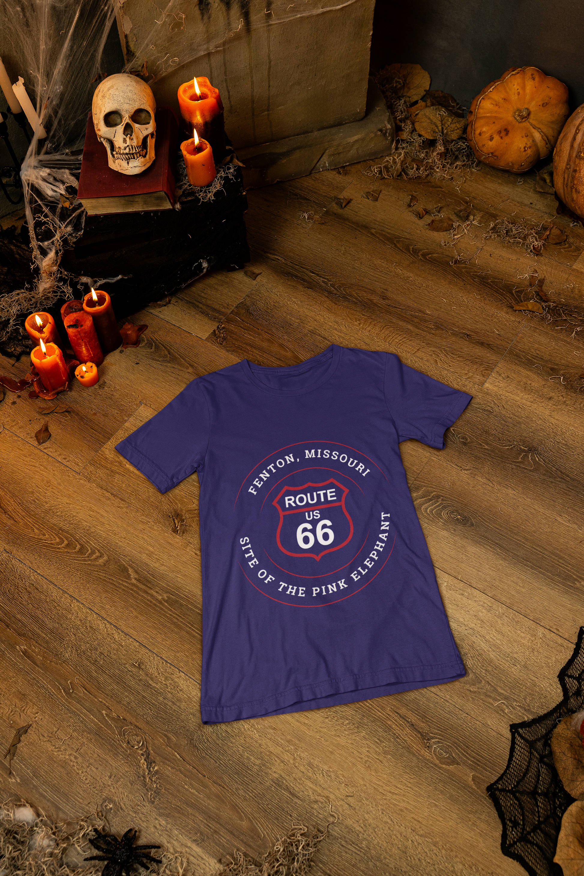 A heather team purple retro Route 66 unisex jersey tee with a Fenton, MO: "Site of the Pink Elephant" design is displayed on the floor with Halloween items such as candles, skulls and candles surrounding it
