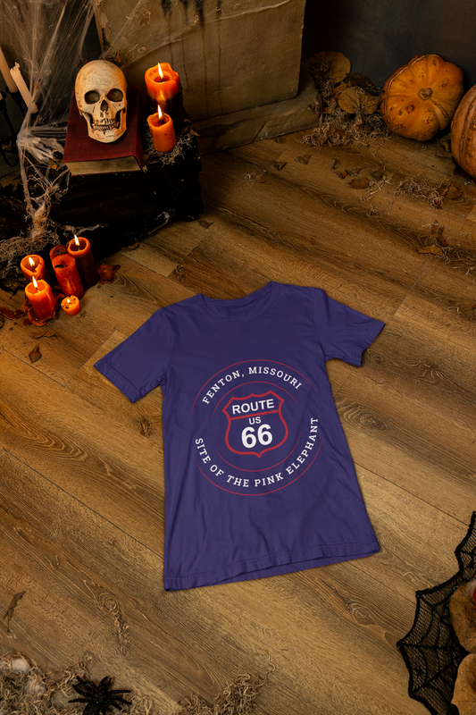 A heather team purple retro Route 66 unisex jersey tee with a Fenton, MO: "Site of the Pink Elephant" design is displayed on the floor with Halloween items such as candles, skulls and candles surrounding it