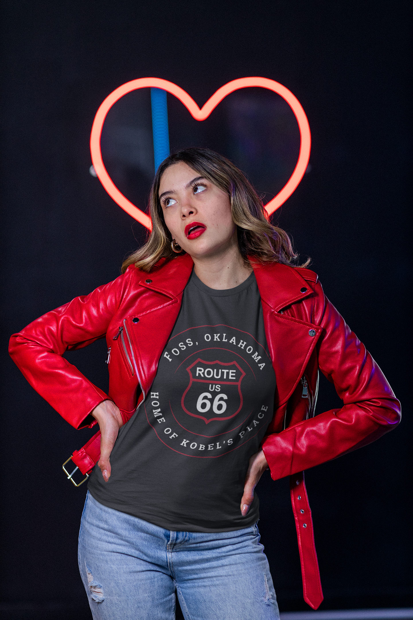 Female model wearing vintage black retro Route 66 unisex jersey tee with Foss, OK "Home of Kobel's Place" design and a red leather jacket with a heart-shaped neon light behind her