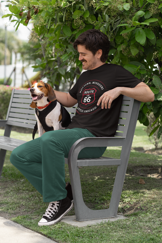 Male model sitting on a bench with his dog. He is wearing a black heather retro Route 66 unisex jersey tee with a Halltown, MO "Little Chicago" design