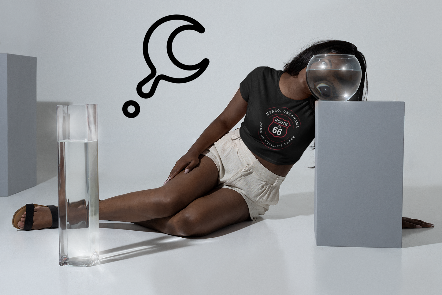 A female model wearing a black heather retro Route 66 unisex jersey tee with Hydro, OK "Home of Lucille's Place" design. She is posed laying on the floor with her face behind a fishbowl, magnifying her eyes for a stylistic shot.