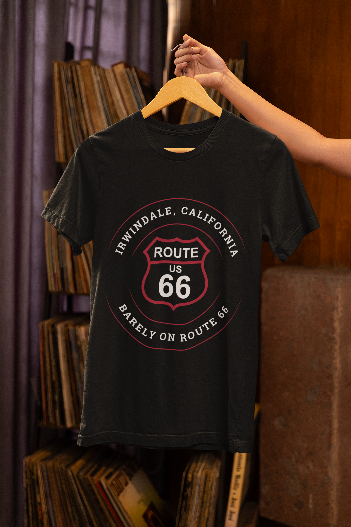 A hand is holding a black retro Route 66 unisex jersey tee with Irwindale, CA: "Barely on Route 66" design hanging on a wooden hanger