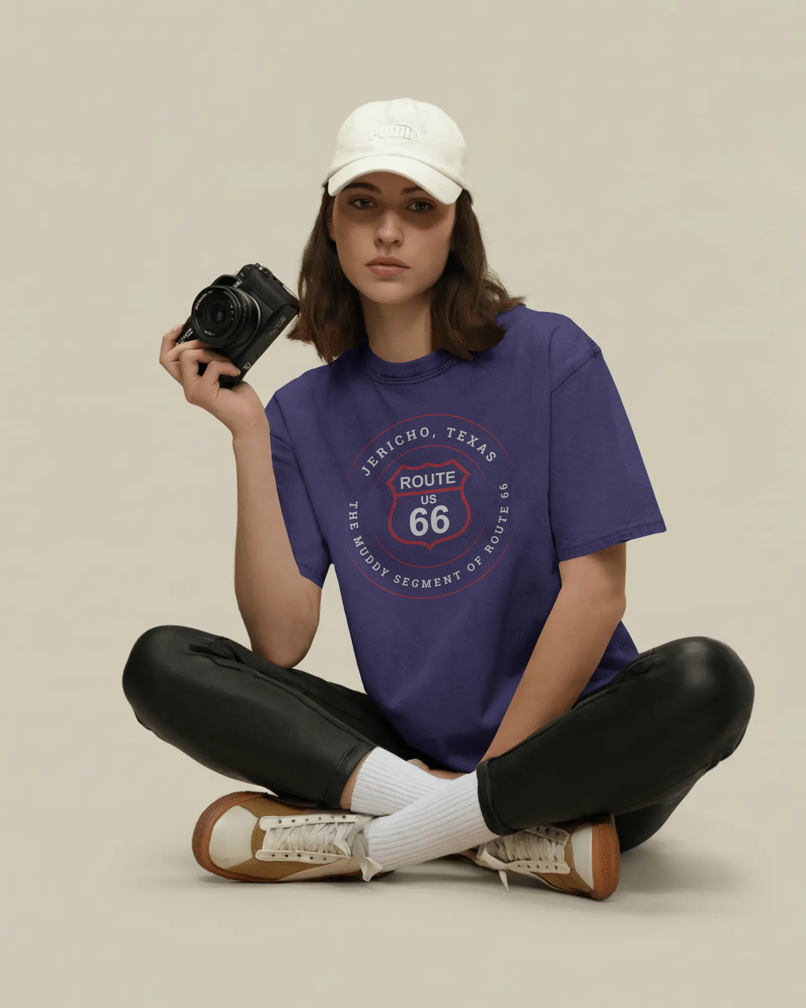 Young woman wearing a team navy retro Route 66 unisex jersey tee with Jericho, TX: The Muddy Segment of Route 66 design, and holding a camera on a beige background