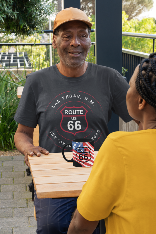 Man wearing black heather retro Route 66 unisex jersey tee with Las Vegas, NM: "The Other Las Vegas" design talks with another person while drinking coffee from a Route 66 cup with a US flag design