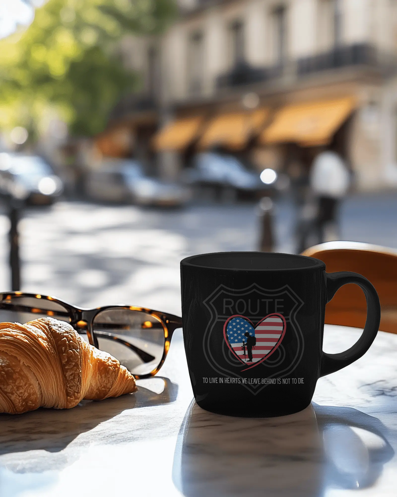 A cafe table which has a croissant, eyeglasses and a black Route 66 latte mug with a heart-shaped US flag, a Route 66 shield and a Thomas Campbell quote on top of it.