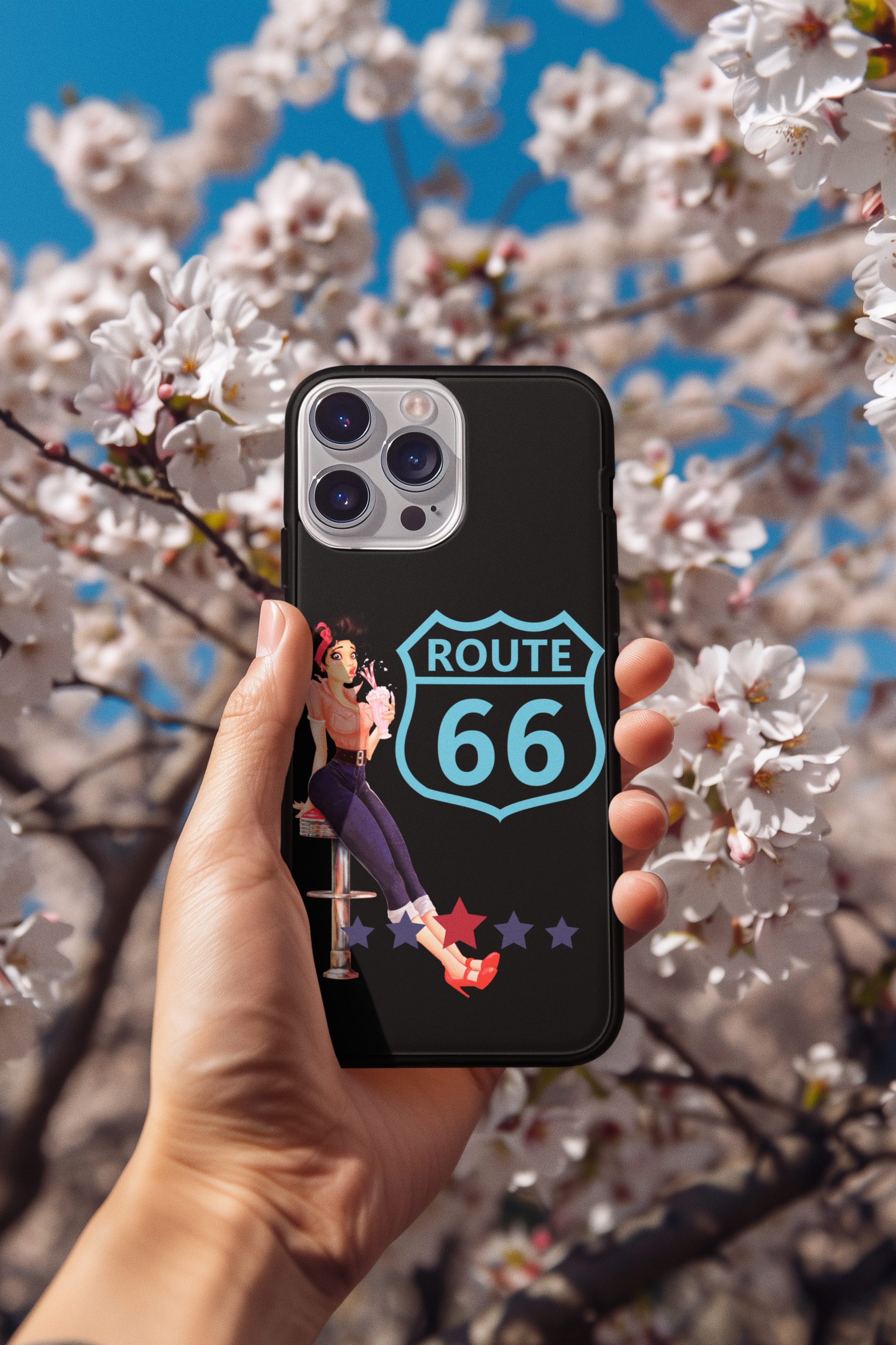 Hand holding Route 66 Milkshake Girl iPhone case against blooming white spring blossoms, showcasing retro diner-inspired phone case design