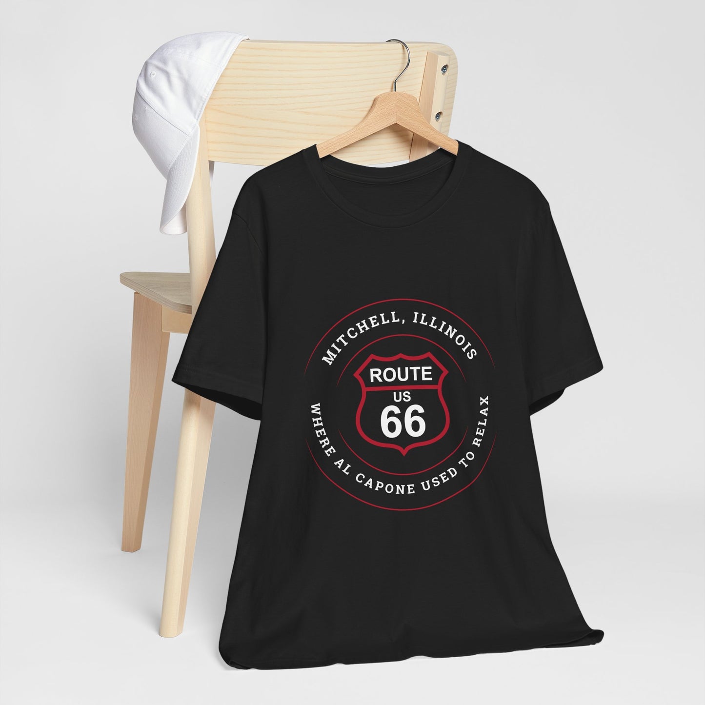 A black retro Route 66 unisex jersey tee with a Mitchell, IL:: "Where Al Capone Used to Relax" design is displayed with a hanger which is hooked to a chair