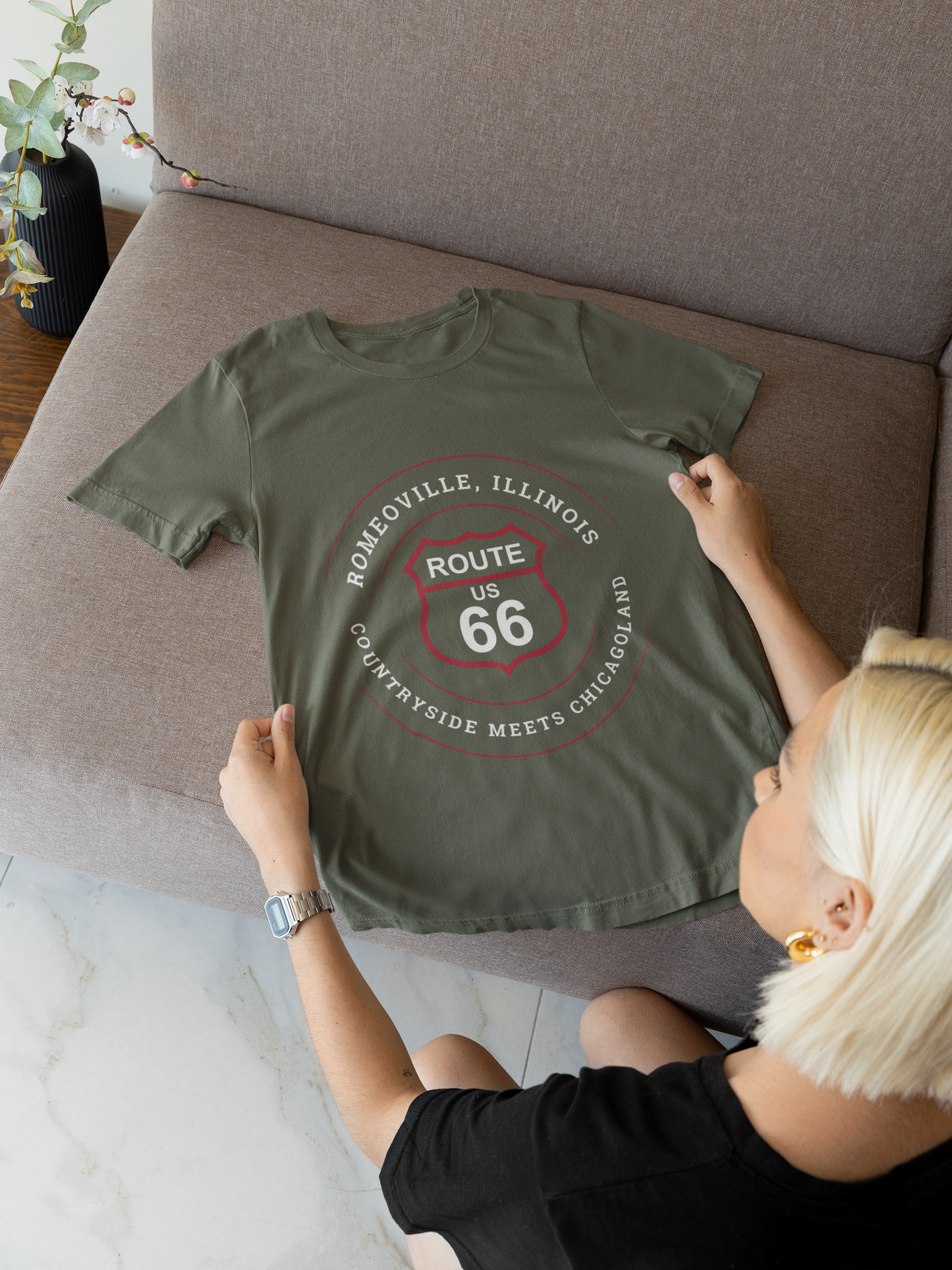 A heather military green retro Route 66 unisex jersey tee. The t-shirt is displayed on a couch. The graphic is a Romeoville, IL: "Countryside Meets Chicagoland" design 