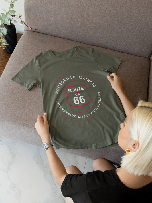 A heather military green retro Route 66 unisex jersey tee. The t-shirt is displayed on a couch. The graphic is a Romeoville, IL: "Countryside Meets Chicagoland" design 