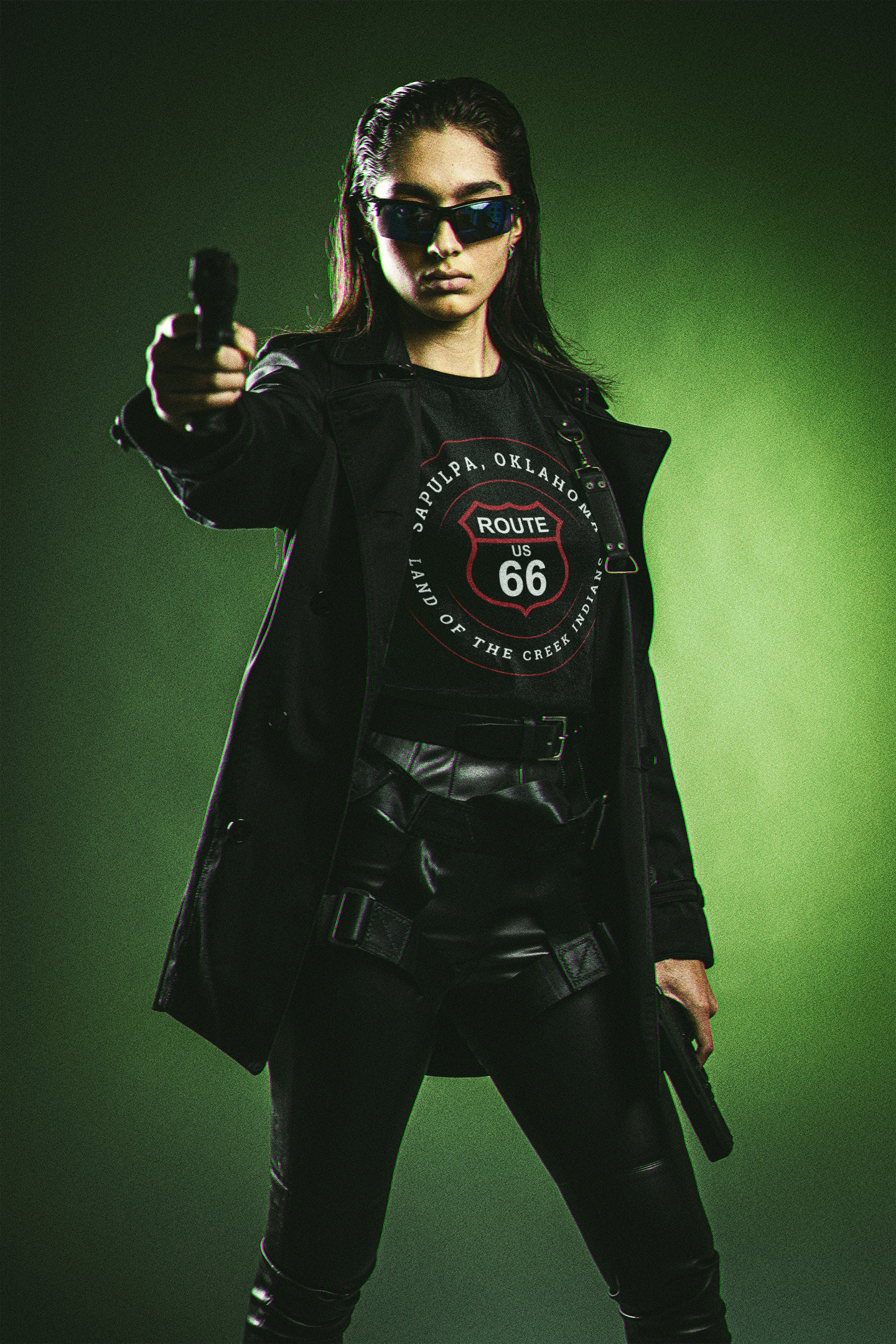 Female model in black leather pants, a trench coat, and black sunglasses, holding a gun while wearing a black retro Route 66 unisex jersey tee with Sapulpa, OK "Land of the Creek Indians" design