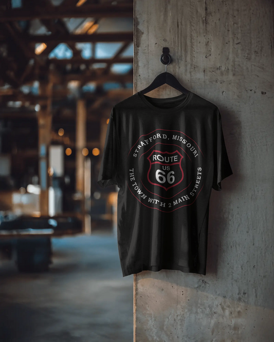 A vintage black retro Route 66 unisex jersey tee with a Strafford, MO "The Town With 2 Main Streets" design hanging on a black hanger against a concrete wall
