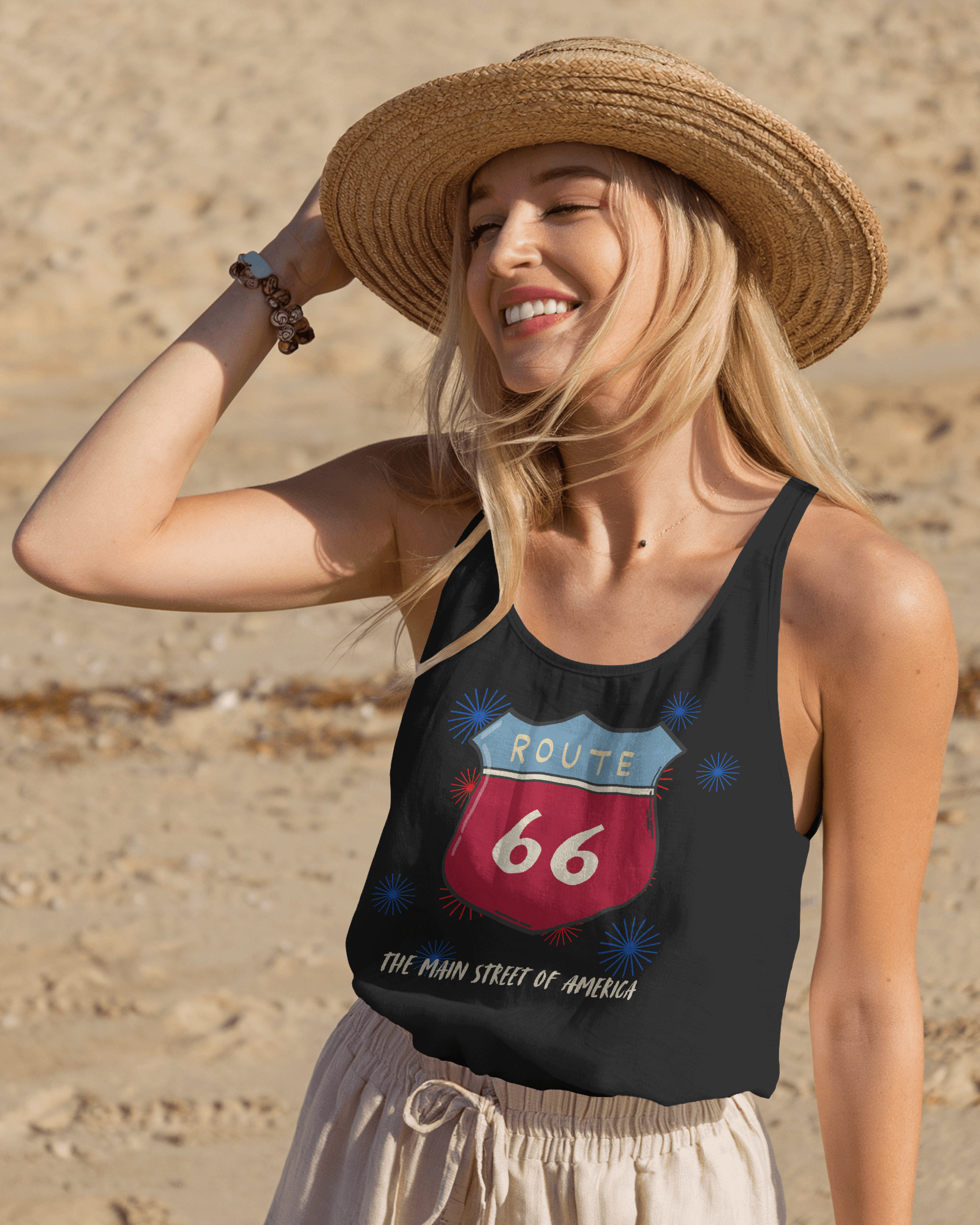 A smiling woman with blonde hair wearing a straw hat and a black tank top is standing on a sandy beach. The tank top features a classic red and blue Route 66 shield sign graphic surrounded by fireworks-like bursts, with the text "THE MAIN STREET OF AMERICA" underneath.