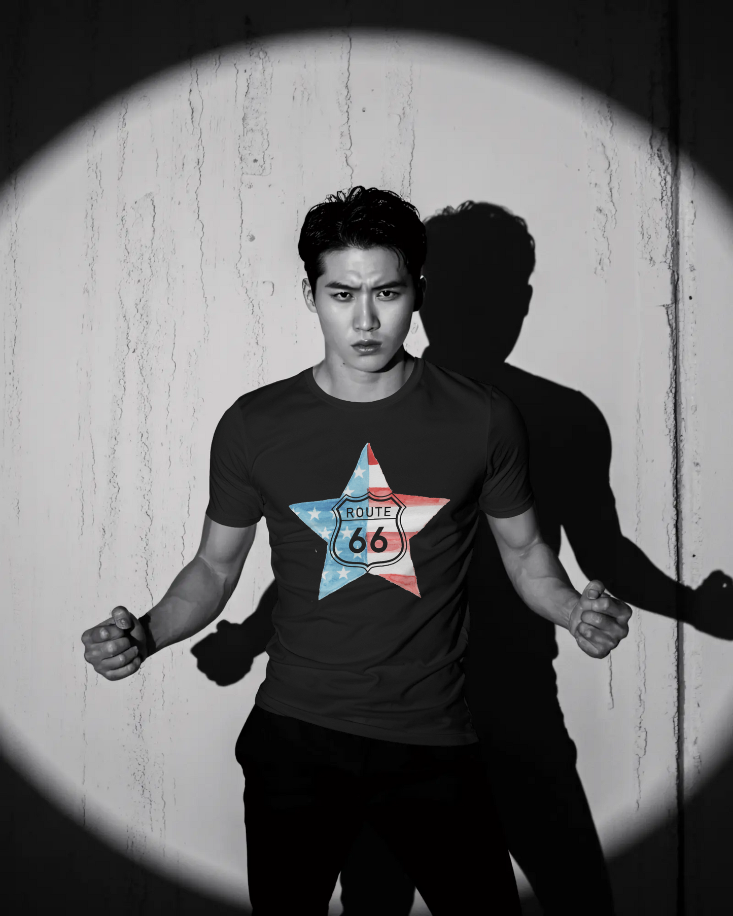 A dramatic, black and white photo of a man posing in a dark t-shirt, highlighted by a spotlight on a textured wall, casting a sharp shadow. The shirt's graphic is a star-shaped American flag with the Route 66 shield logo in the center.