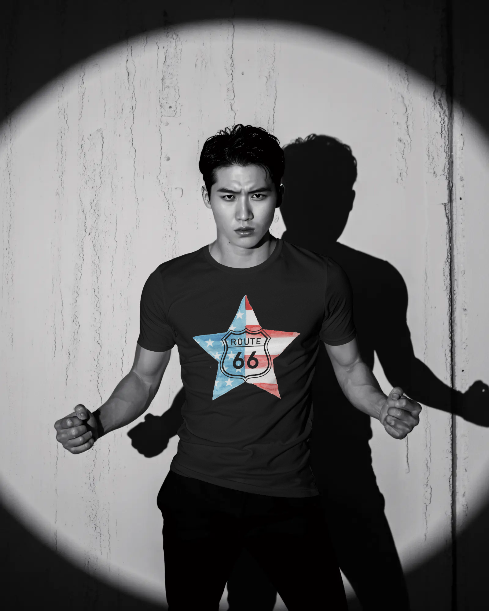 A dramatic, black and white photo of a man posing in a dark t-shirt, highlighted by a spotlight on a textured wall, casting a sharp shadow. The shirt's graphic is a star-shaped American flag with the Route 66 shield logo in the center.