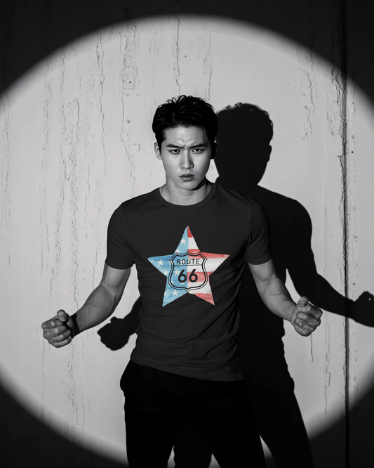A dramatic, black and white photo of a man posing in a dark t-shirt, highlighted by a spotlight on a textured wall, casting a sharp shadow. The shirt's graphic is a star-shaped American flag with the Route 66 shield logo in the center.