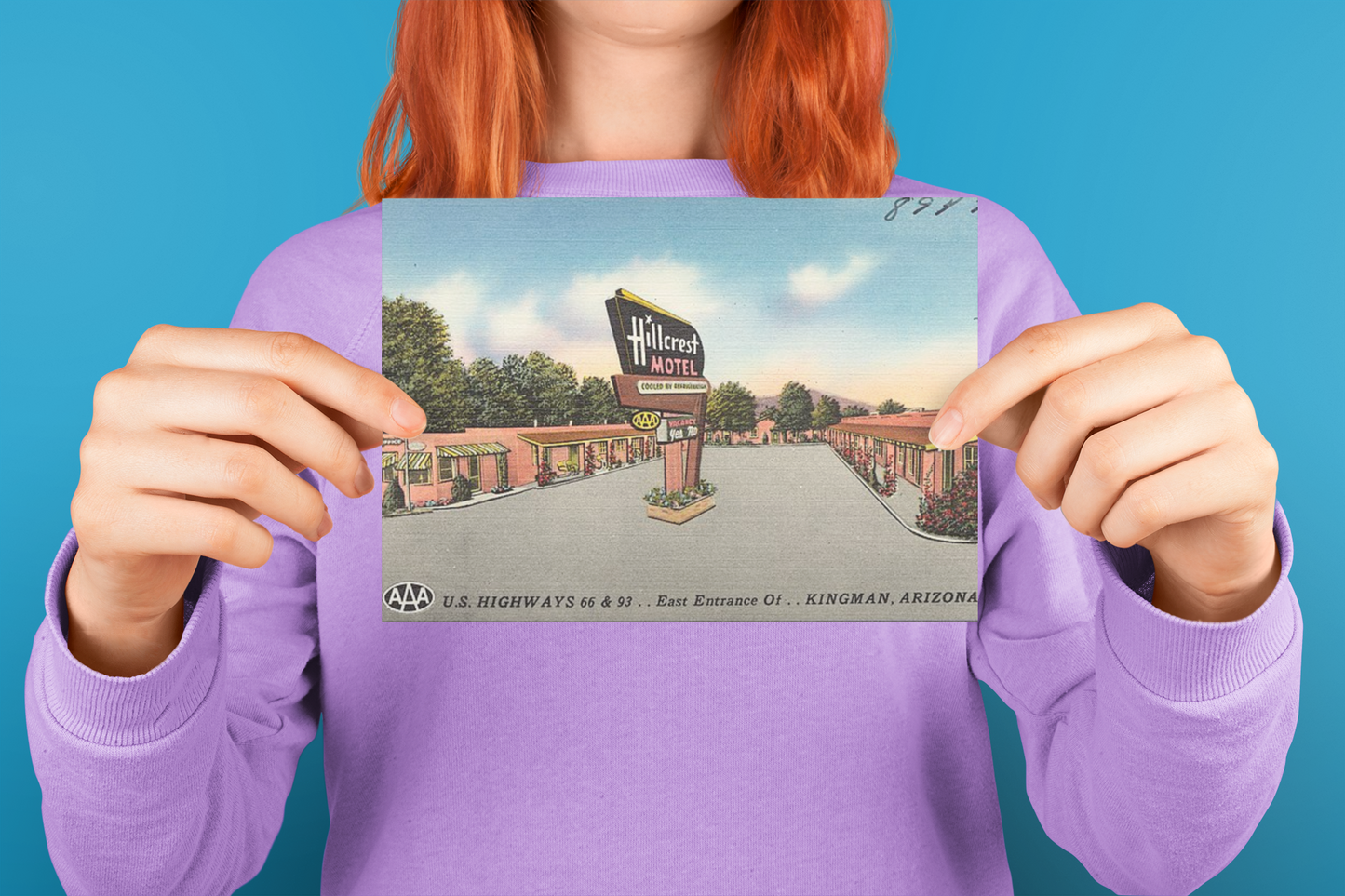 A woman with red hair holding a vintage Hillcrest Motel postcard from Kingman, Arizona showing the front illustration of the motel sign, building and a Route 66 classic scene