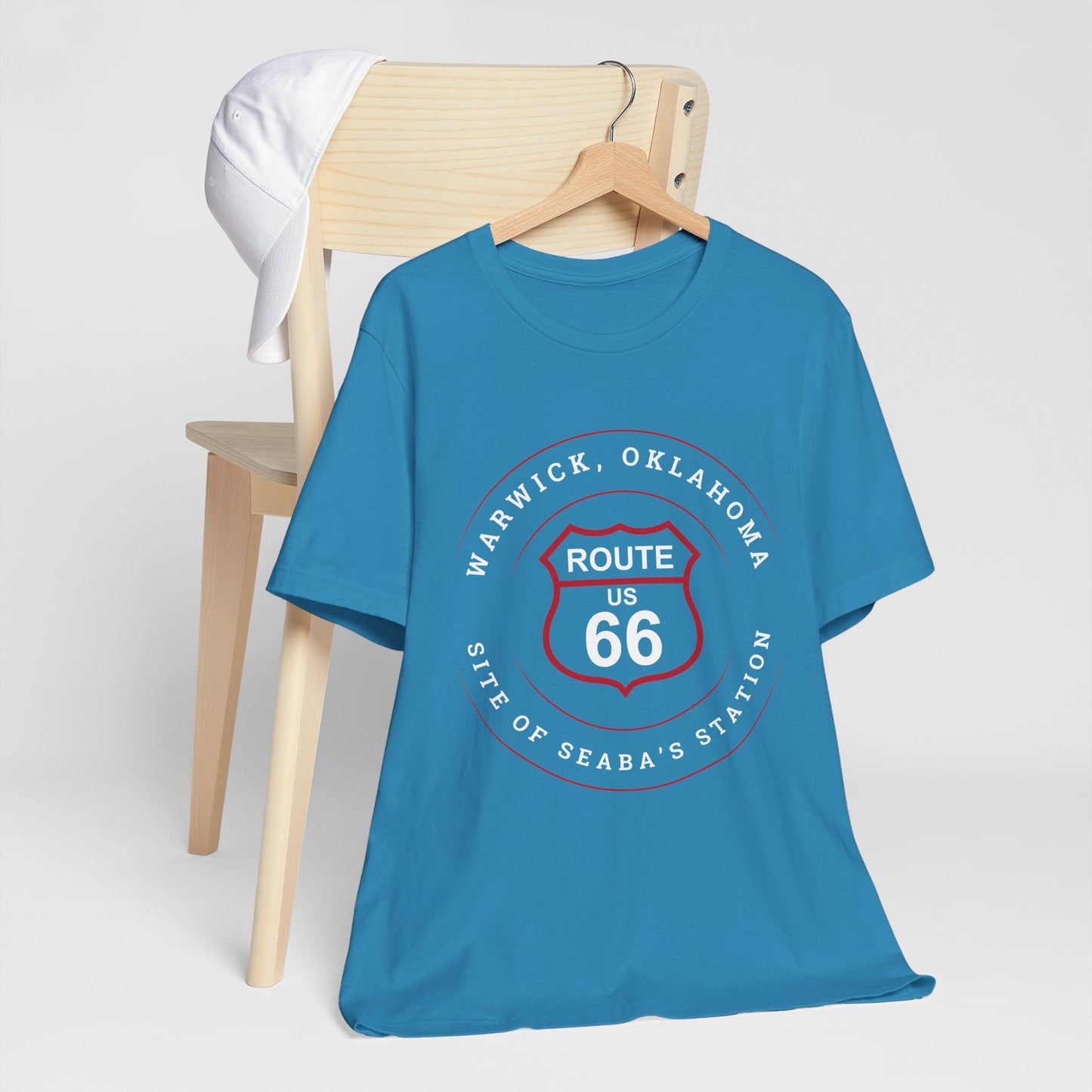 Aqua retro Route 66 unisex jersey tee with Warwick, OK "Site of Seaba's Station" design hanging on a wooden hanger. It is hanging of a wooden chair with the bottom gently grazing the floor.