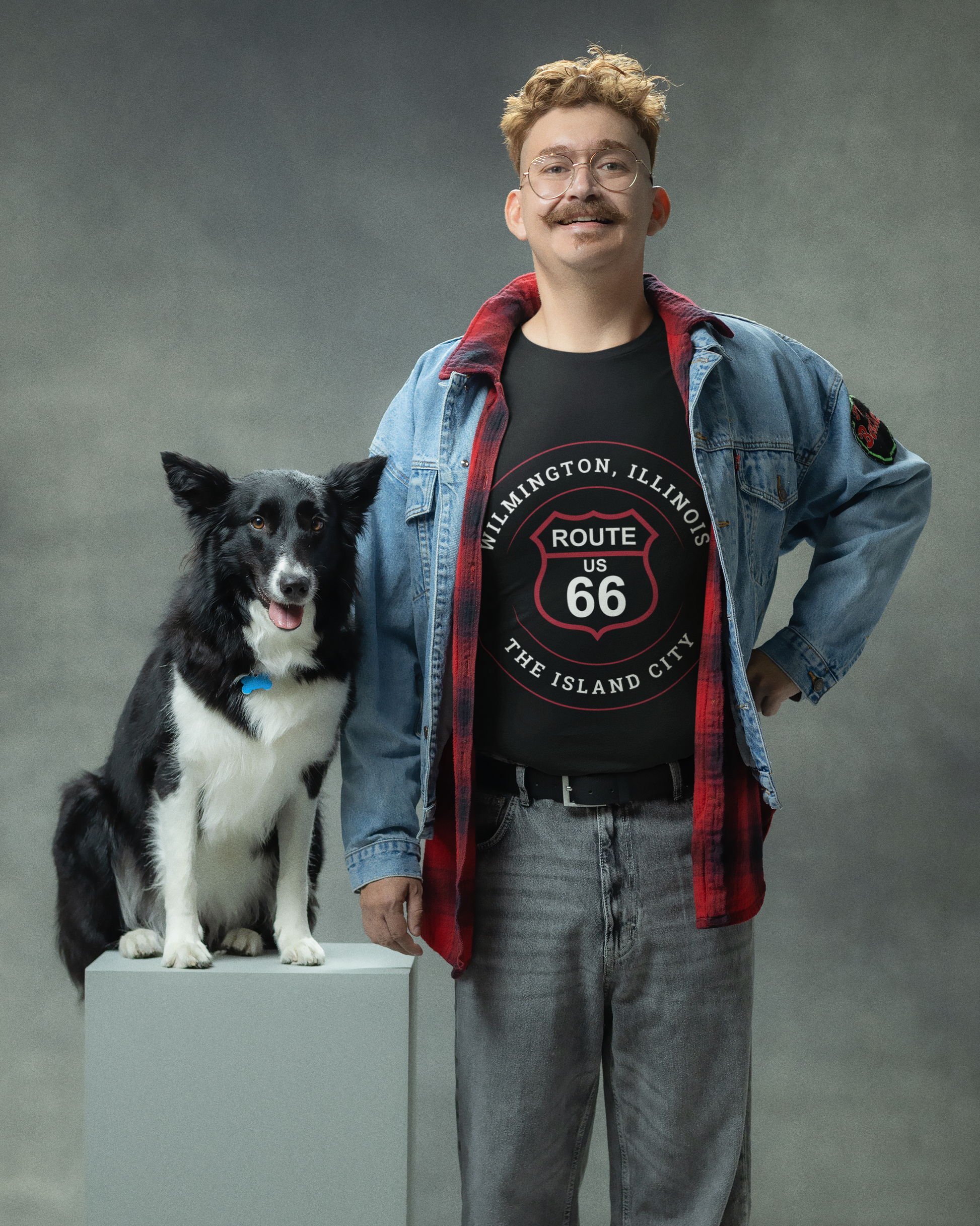 Male model standing next to his dog and he is wearing a black heather retro Route 66 unisex jersey tee with a Wilmington, IL: "The Island City" design