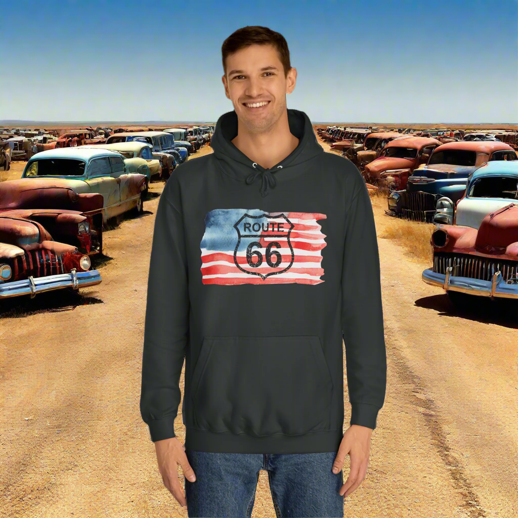Man smiling and modeling a jet black Route 66 hoodie with a patriotic watercolor-styled American flag design across the chest. He is standing at a Route 66 lot which has multiple classic cars.